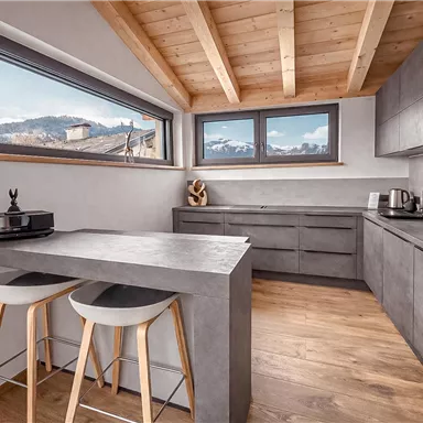 Eine moderne Küche mit Holzdecken und großen Fenstern. Der Raum ist mit grauen Möbeln und einem Tisch mit Hockern ausgestattet.