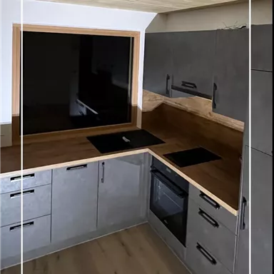 Eine moderne Küche mit grauen Schränken und einer Holzarbeitsplatte. Der Raum hat große Fenster und eine helle Atmosphäre.