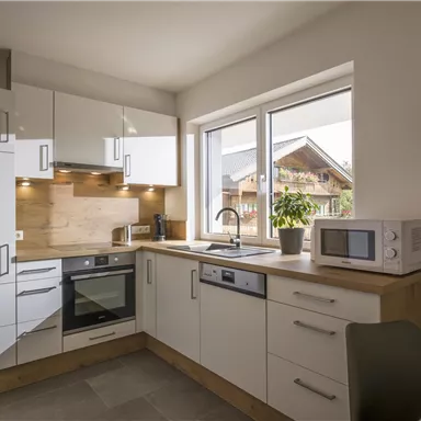 A modern kitchen with white cabinets and wooden details. There is plenty of daylight at the window, and the facilities include an oven and a microwave.