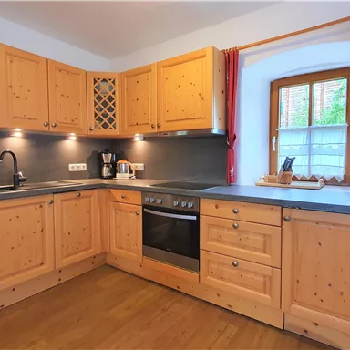 Eine moderne Küche mit Holzschränken und dunkler Arbeitsplatte. Es gibt eine Spüle, einen Herd und Fenster, die Licht hereinlassen.