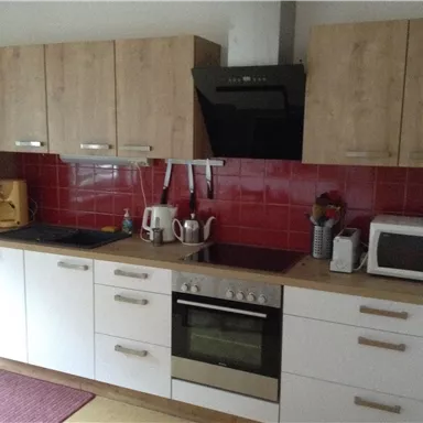 A modern kitchen with wooden cabinets and a red tiled wall. On the countertop are kitchen appliances like a kettle and a microwave.