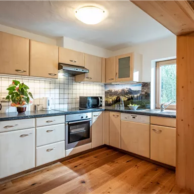 A modern kitchen with wooden cabinets and a large window. In the foreground, there is a plant and a refrigerator is visible.