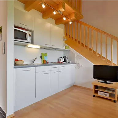 Eine moderne Küche mit hellen Schränken und einem Kühlschrank. Im Hintergrund sieht man eine gemütliche Sitzecke und eine Treppe.