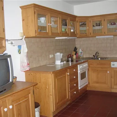 A modern kitchen with wooden cabinets and a bright countertop. In the background, cooking utensils and a television are visible.