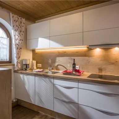 A modern kitchen with white cabinets and wooden surfaces. The light pleasantly brightens the room through a window with curtains.