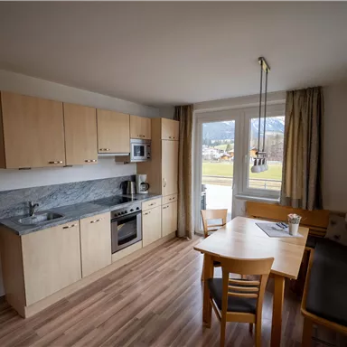 Eine helle Küche mit Holzschränken und modernen Geräten. Ein Esstisch mit Stühlen steht in der Ecke und bietet einen Blick nach draußen.