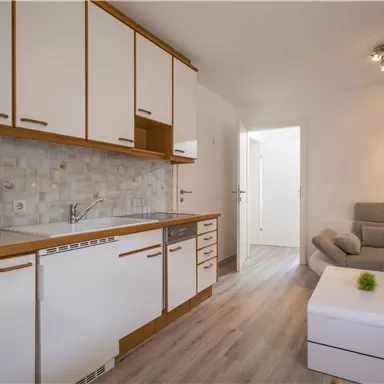 A modern kitchen corner with white cabinets and a cozy living room. The room is bright and inviting, with a gray sofa and a small table.