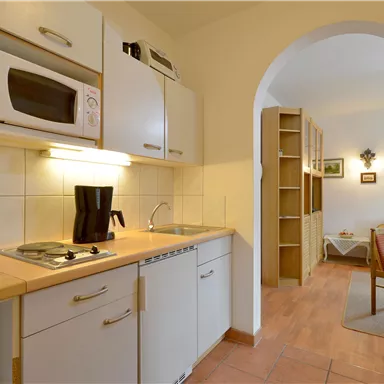 Eine kleine, moderne Küche mit weißen Schränken und einer Ausstattung wie Mikrowelle und Wasserkocher. Im Hintergrund ist ein gemütlicher Wohnbereich sichtbar.