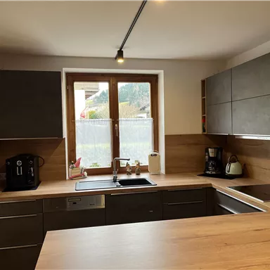 Eine moderne Küche mit Holzoberflächen und grauen Schränken. Es gibt ein Fenster mit Blick ins Freie und eine Kaffeemaschine auf der Arbeitsfläche.