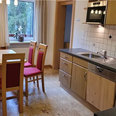 Eine moderne Küche mit einer hellen Ausstattung und einem angrenzenden Esstisch. Die Möbel sind in warmen Holz- und Rottönen gehalten.