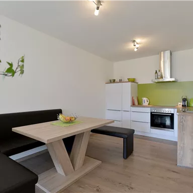 A modern kitchen with a wooden dining table and an elegant bench. The walls are in light green and white, creating a friendly atmosphere.