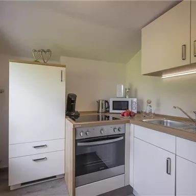 Eine moderne Küche mit weißen Schränken und einem Herd. Auf der Arbeitsfläche stehen Küchenutensilien und frisches Obst.