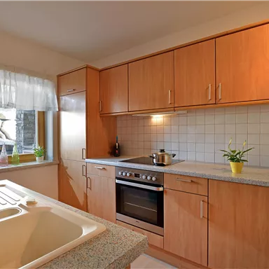 A modern kitchen with wooden cabinets and a bright countertop. A cooking pot is on the stove, and there are windows with a view of the outdoors.