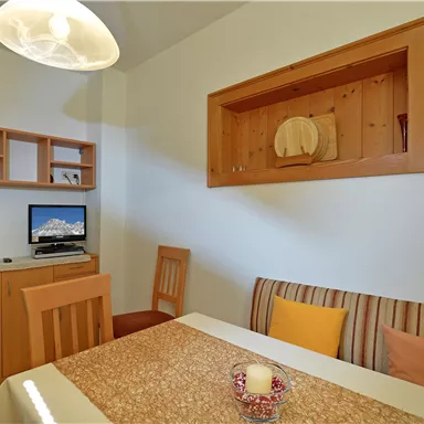 Ein gemütlicher Essbereich mit einem Tisch, Stühlen und einer Sofa-Ecke. An der Wand befindet sich ein Regalsystem und ein Fernseher.