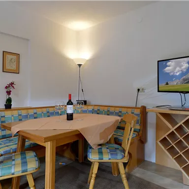 Ein gemütlicher Wohnbereich mit einem Esstisch und bunten Stühlen. An der Wand befindet sich ein Fernseher und eine Flasche Wein auf dem Tisch.