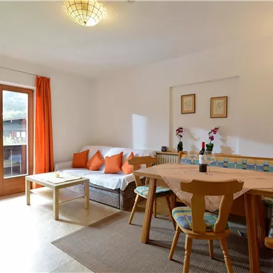 Ein gemütlicher Wohnraum mit orangefarbenen Vorhängen und einer Essecke. Es gibt ein Sofa mit Kissen und einen Tisch in der Mitte.