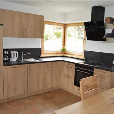 Eine moderne Küche mit Holzschränken und schwarzer Arbeitsplatte. Viel natürliches Licht durch große Fenster.