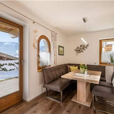 An inviting dining area with a large table and a cozy seating corner. Through the windows, one can see a snow-covered landscape.