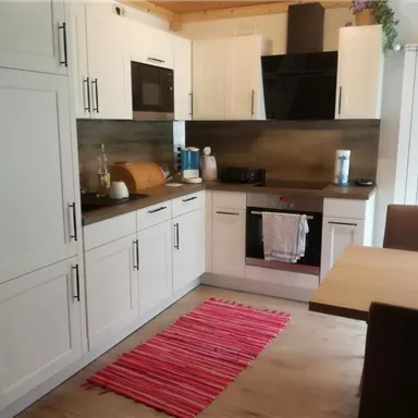 A modern kitchen with white cabinets and a wooden countertop. The table is made of wood, and a red carpet is on the floor.