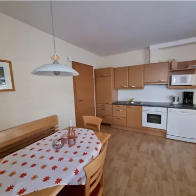 Eine moderne Küche mit Holzschränken und einem Esstisch. Auf dem Tisch liegt eine rot-weiße Tischdecke und eine Flasche Wasser.