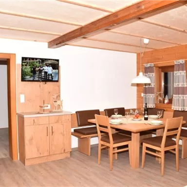 A cozy dining area with a wooden table and chairs. In the background, a kitchen and a television are visible.