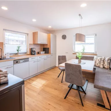 Eine moderne Küche mit Holzoberflächen und ausreichend Stauraum. Der Essbereich besteht aus einem Tisch und bequemen Sitzen, umgeben von Tageslicht.