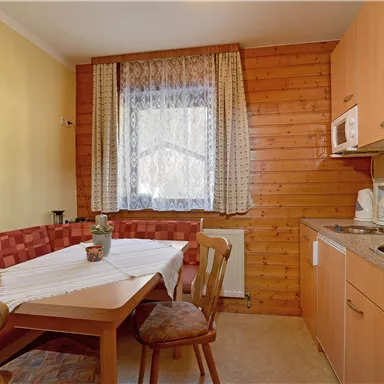 Eine gemütliche Küche mit einem Holztisch und Stühlen. Die Wände sind aus Holz und der Raum hat große Fenster mit Vorhängen.