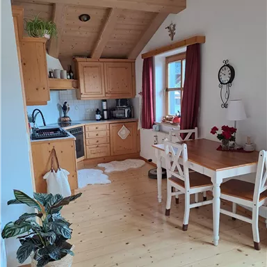 Eine gemütliche Küche mit Holzschränken und einem Esstisch. Pflanzen und rote Vorhänge verleihen dem Raum eine einladende Atmosphäre.