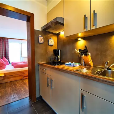 Eine helle Küche mit modernen Geräten und Holzregalen. Im Hintergrund ist ein Schlafzimmer mit einem großen Bett sichtbar.