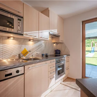 Eine moderne Küche mit Holzschränken und eingebauten Geräten. Durch das Fenster sieht man einen Gartenbereich mit Sitzgelegenheiten.