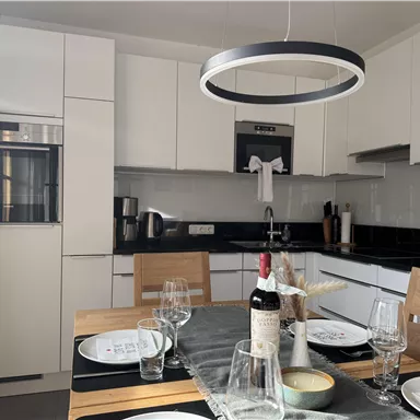 Modern kitchen with elegant white cabinets and a wooden table that is set. In the background, a refrigerator and a stove are visible, along with decorative elements.