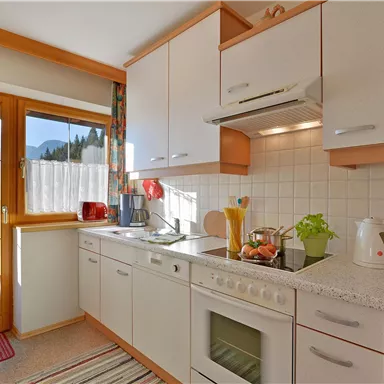 Eine helle Küche mit modernen Geräten und einem großen Fenster, das viel Tageslicht hereinlässt. Auf der Arbeitsfläche sind frisches Obst und Küchenutensilien sichtbar.