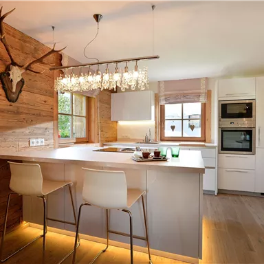 Eine moderne Küche mit Holzverkleidung und eleganten Barhockern. Im Hintergrund sind Fenster und ein stilvoller Kronleuchter zu sehen.