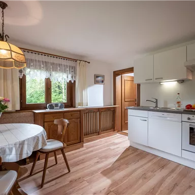 Eine helle Küche mit einem runden Tisch, Stühlen und einer modernen Ausstattung. Große Fenster lassen viel Licht herein und bieten einen Blick ins Grüne.