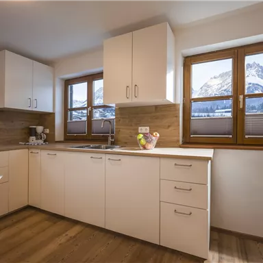 Eine moderne Küche mit weißen Schränken und Holzverkleidung. Große Fenster bieten einen Blick auf die Berge.