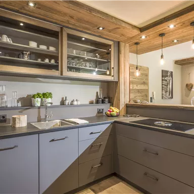 Eine moderne Küche mit grauen Schränken und Holzdetails. Es gibt eine Spüle, einen Kochbereich und helle Beleuchtung.