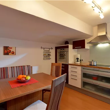 Eine moderne Küche mit einem Esstisch und Stühlen. Die Wände sind hell und dekoriert mit Bildern, während frisches Obst auf dem Tisch liegt.