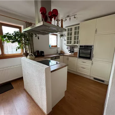 Eine moderne Küche mit hellen Schränken und einem Kochfeld in der Mitte. Große Fenster sorgen für viel Tageslicht und eine grüne Pflanze bringt Frische in den Raum.