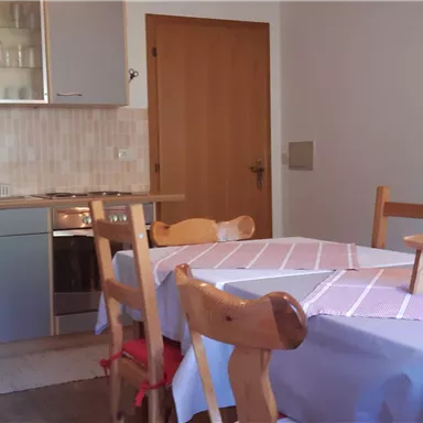 Eine helle Küche mit einem Holztisch und Stühlen. Im Hintergrund sind eine Spüle und Küchenschränke zu sehen.