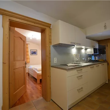 A modern kitchen with wooden details and a bright entrance area. In the background, a bedroom can be seen.