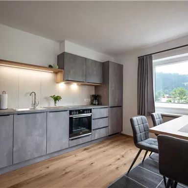 A modern kitchen with gray cabinets and a dining table. Large windows provide a view of the surroundings and ensure plenty of daylight.