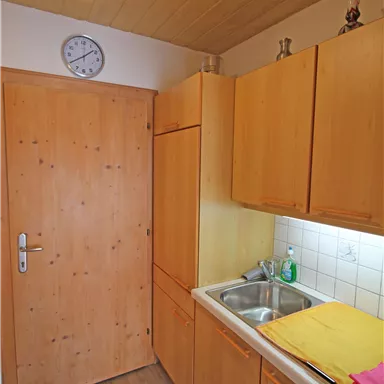 Eine kleine Küche mit Holzschränken und einem Waschbecken. An der Wand hängt eine Uhr und es liegen bunte Handtücher auf der Arbeitsfläche.