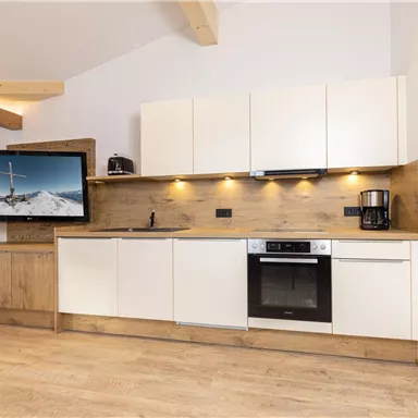 Eine moderne Küche mit hellen Schränken und Holzakzenten. Ein Fernseher hängt an der Wand, und die Ausstattung ist funktional und stilvoll.