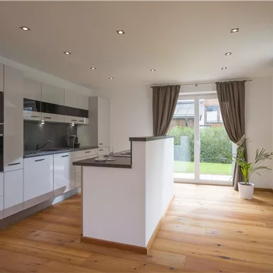 Eine moderne Küche mit weißen Schränken und Holzfußboden. Große Fenster bringen viel Licht und bieten einen Blick auf den Garten.