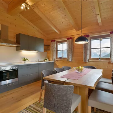 A modern wooden kitchen with a dining table and chairs. Large windows provide plenty of daylight and a view outside.