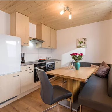 A modern kitchen unit with wooden cabinets and a dining table. The decor is bright and friendly, with a bouquet of flowers on the table.