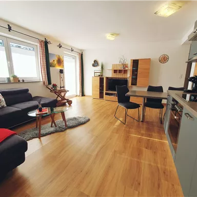 Ein gemütlicher Wohnraum mit einem schwarzen Sofa und einem Holztisch. Große Fenster sorgen für viel Tageslicht.