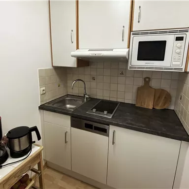 Eine kleine Küche mit einer Spüle, einem Elektroherd und einer Mikrowelle. Neben der Küchenzeile stehen eine Kaffeekanne und ein Holzbrett.