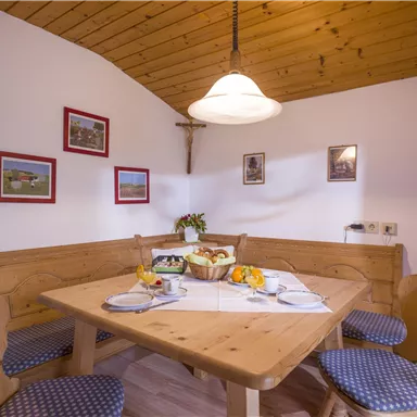 Ein gemütlicher Essbereich mit Holzmöbeln und blauen Sitzkissen. An den Wänden hängen Bilder und es steht ein Obstkorb auf dem Tisch.