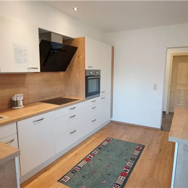 Eine moderne Küche mit Holzoberflächen und weißen Schränken. Der Raum ist hell und offen mit einem Teppich in der Mitte.